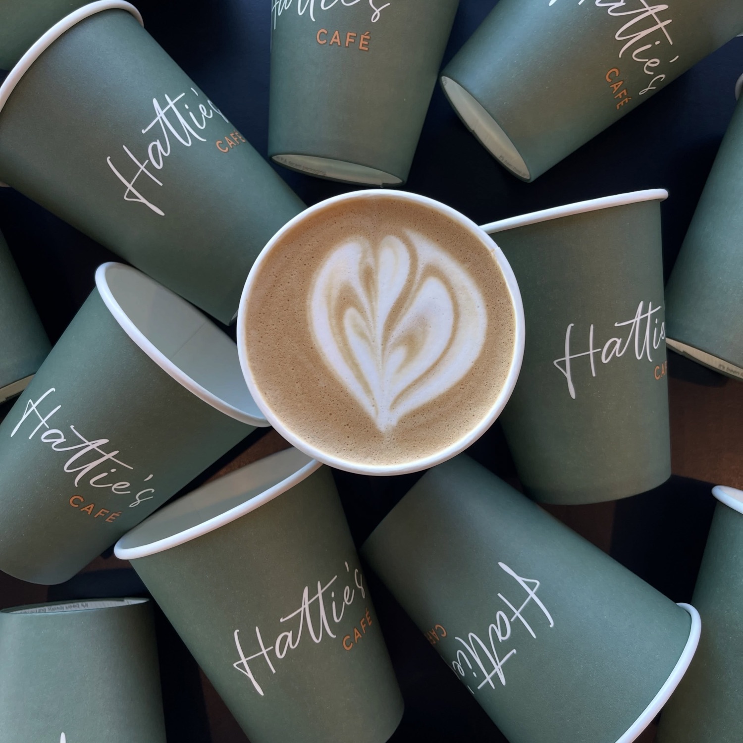 A latte with leaf art surrounded by green Hattie's Cafe takeaway cups, seen from above